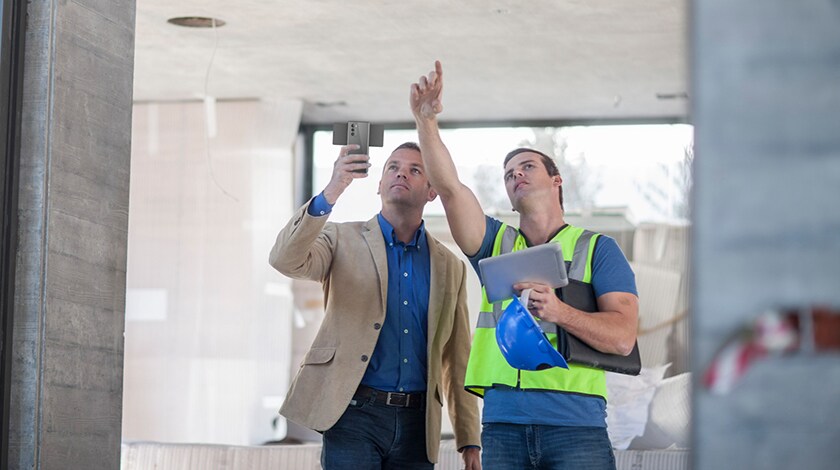 Two men point up to something construction-related and hold a smartphone and tablet.