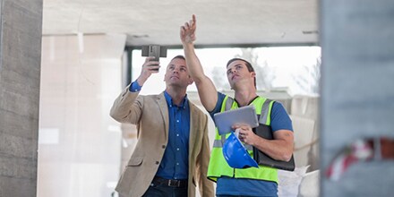 Two men point up to something construction-related and hold a smartphone and tablet.