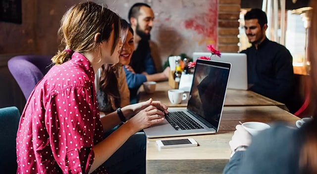 Two smiling women at a desk view a common laptop screen. In the background, two men collaborate on a single laptop, too.