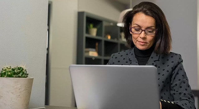 A brunette woman in black glasses and a grey blazer works on a laptop.