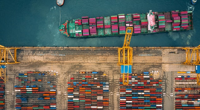 An overhead view of brightly colored shipping containers being unloaded by a yellow crane at a port.