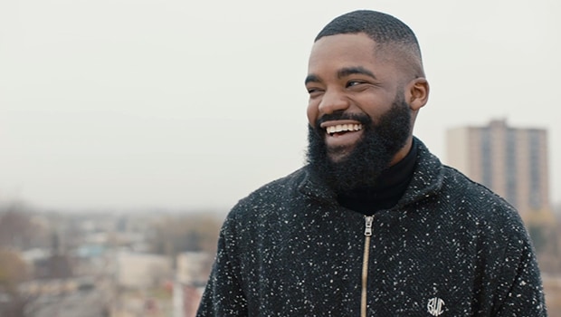 Will Toms wears black hoodie, smiling and looking to the side with a blurred-out city behind him.
