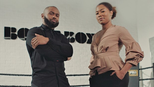 Man in all black poses beside a woman, as both stare at the camera.