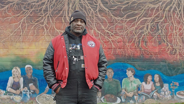 Man in winter hat and clothing stands outside in front of a colorful mural.