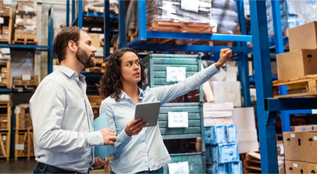 Two warehouse workers gesture to shelves while one holds a tablet.