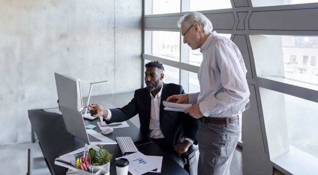 Two government employees collaborate over multiple computers at the office.