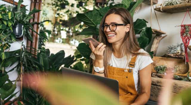 A worker at a plant shop sits and smiles while talking on their smartphone.