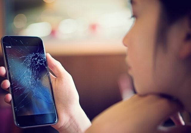A young woman looks at a smartphone with a cracked screen.