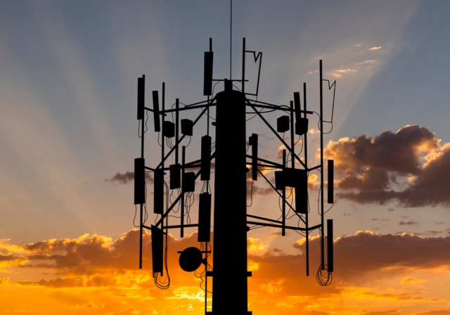 Top of a cell phone tower with multiple signal antennas in front of a sunset at dusk.