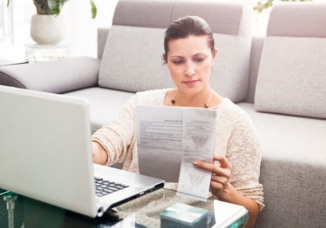 Woman using a laptop and looking at a document she is holding.