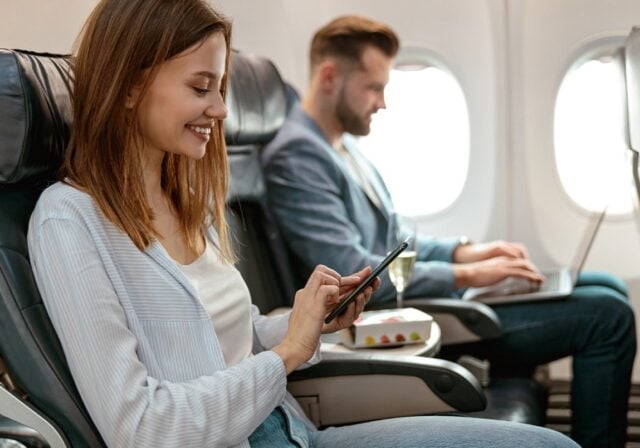 A smiling woman on an airplane scrolling on her phone while sitting next to a man typing on his laptop.