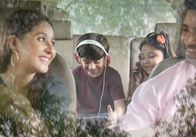 A happy family of four riding in the car with two kids in the back seat watching a tablet.