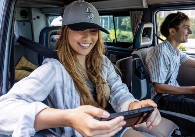 A young woman in the passenger seat of an RV watches her smartphone while a young man drives.