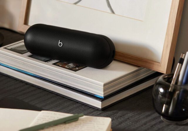 A black Beats portable speaker resting on top of stacked books near a framed picture, with a pen holder and notebook on a desk.