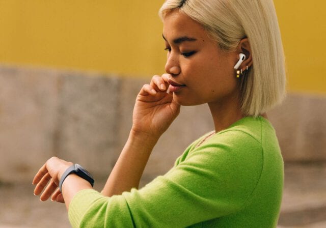 Woman wearing wireless earbuds and a smartwatch looking at her wrist outdoors.