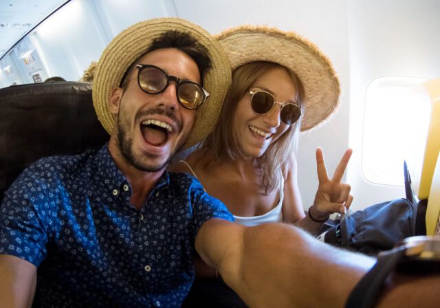 Two happy travelers wearing straw hats and sunglasses take a cheerful selfie on an airplane. The man is smiling widely with his mouth open, and the woman beside him is making a peace sign. Sunlight shines through the airplane window behind them.