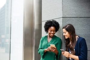 Two women standing outside smiling and looking at smartphones together.