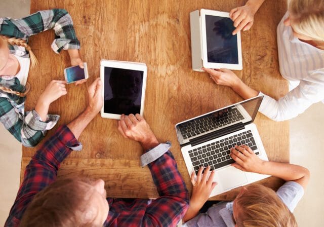 Family sitting around a wooden table using a smartphone, tablets, and a laptop.