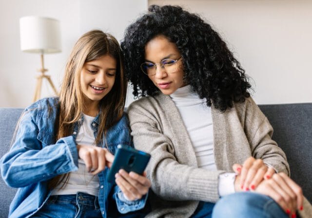Woman and girl smiling while looking at a smartphone together on a couch.