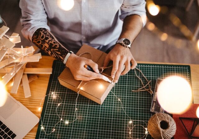 A person with a tattooed arm wraps a gift box with twine at a crafting table decorated with string lights, a laptop, and holiday materials.