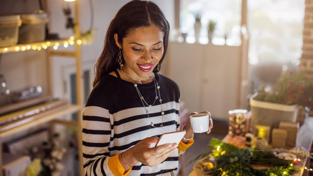 A woman in a cozy, warmly lit room smiles while looking at her smartphone and holding a small cup, surrounded by holiday décor and gift-wrapping supplies.