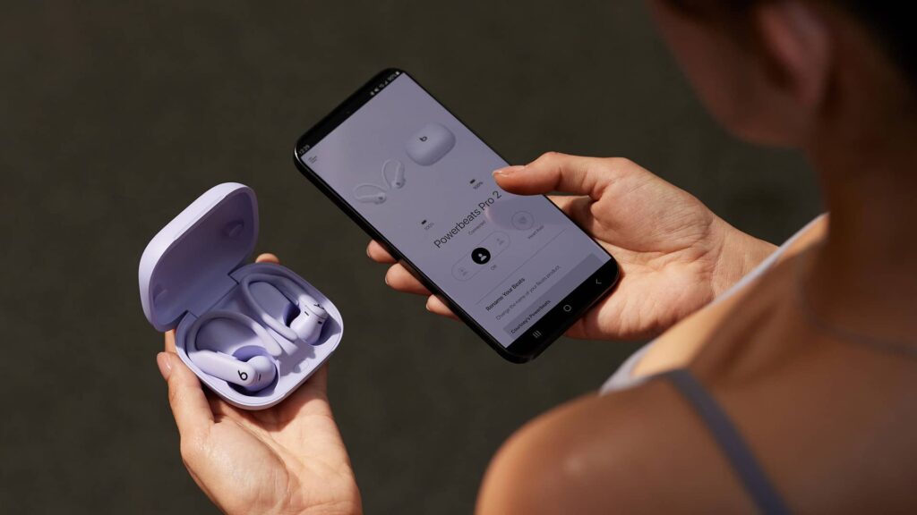 A person holds an open lavender-colored case containing Powerbeats Pro 2 earbuds in one hand while using a smartphone in the other hand to view the device’s connection and settings screen.