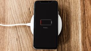 Smartphone lying face up on a wooden surface, placed on a wireless charging pad, with the screen showing a nearly empty battery and the message ‘Charging…’.