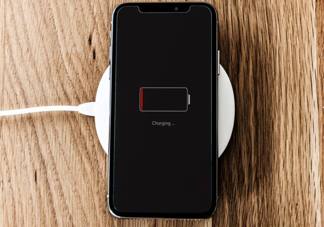Smartphone lying face up on a wooden surface, placed on a wireless charging pad, with the screen showing a nearly empty battery and the message ‘Charging…’.