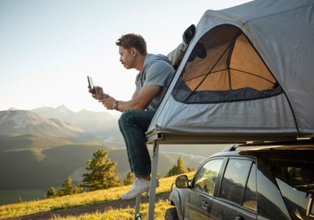 Man sits on the edge of his car tent while taking pictures of a green hilly landscape.