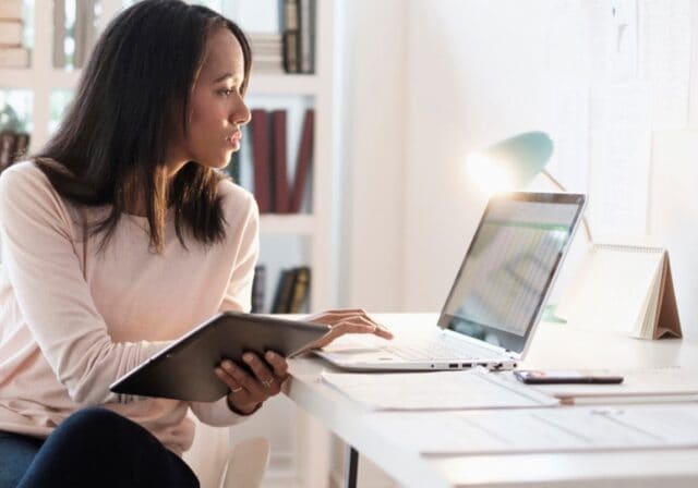 A person looking at their laptop while holding a tablet.