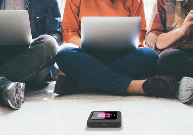 Three individuals sitting down with laptops and phones connecting to the hotspot device in front of them