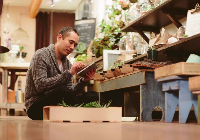 A person sat on the floor using a tablet surrounded by plants.