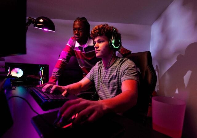 Two men standing at a computer with one engaged with a video game.