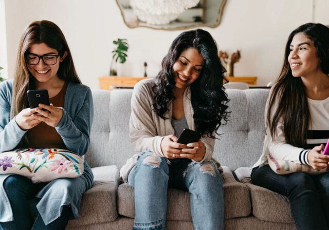 Three people sitting on a couch indoors, each holding a smartphone, with a decorative pillow and plants visible in the background.