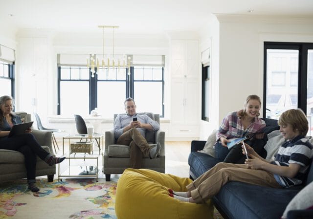 A family of four relaxes together in a bright, modern living room. A woman sits on the left in an armchair using a tablet, while a man in another armchair looks at his smartphone. On the right, a young woman and a boy sit on a dark blue couch, smiling and looking at a tablet. The room is well-lit with large windows, white walls, a colorful rug, and a yellow beanbag on the floor.
