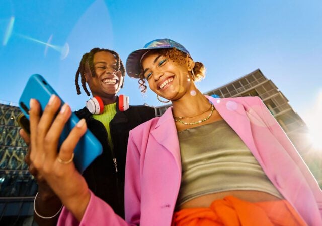 Two people outdoors holding a smartphone, wearing colorful outfits and accessories, with modern glass buildings and a bright blue sky in the background.