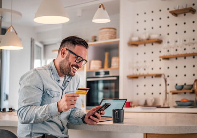 Smiling man sitting in a modern kitchen making an online purchase with a credit card and smartphone, laptop open on the counter beside a coffee mug.
