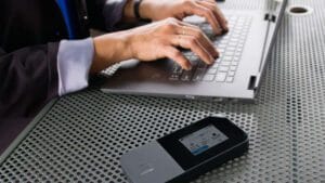 A person typing on a laptop beside a portable Wi-Fi hotspot on a metal table.