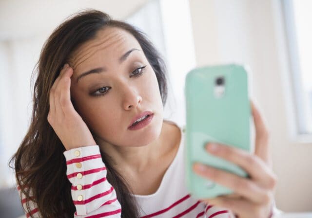 A woman holding a smartphone looks at the screen with a worried or confused expression, touching her forehead with her other hand.