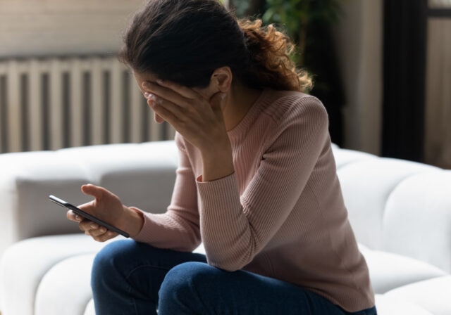 A woman sitting on a couch holds a smartphone in one hand while covering her face with the other, appearing upset or stressed.