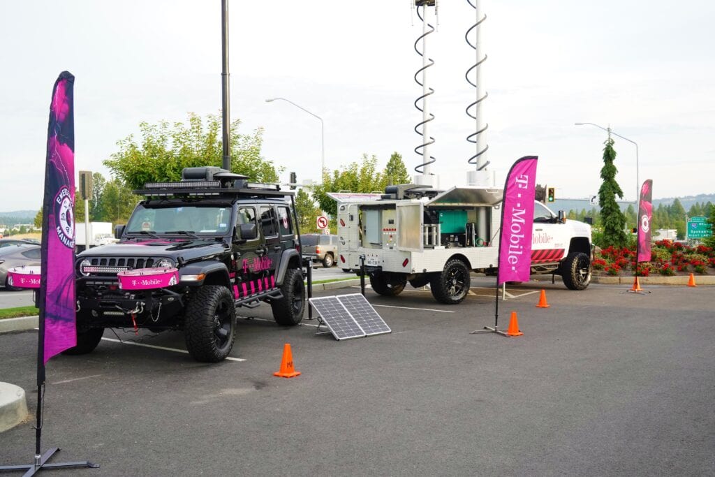 Two heavy duty vehicles parked back-to-back outdoors, branded with the T-Mobile logo. Vehicle on the right of the image has satellite equipment extended