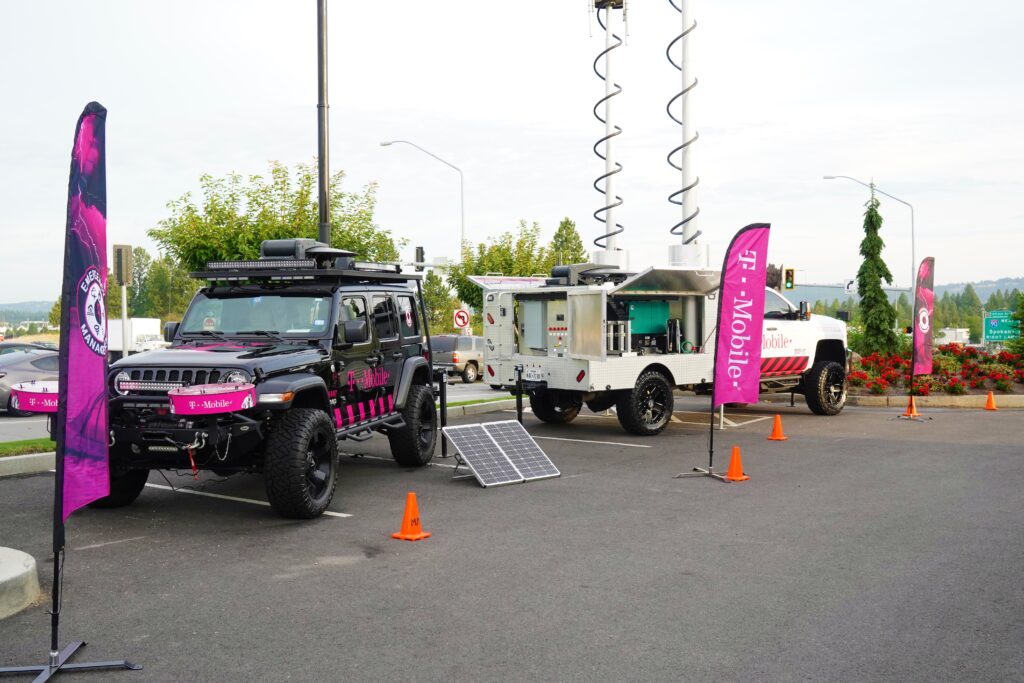 Dos vehículos de uso intensivo estacionados al aire libre uno al lado del otro, con el logotipo de T-Mobile. El equipo satelital del vehículo a la derecha de la imagen está extendido