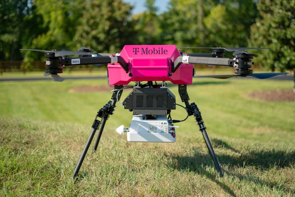 Foto en primer plano de un dron colocado sobre el césped. El dron tiene cuatro hélices y tiene el logotipo de T-Mobile en la parte frontal y central