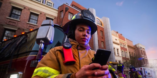 Female firefighter looking at her cell phone in front of a firetruck and brick building.