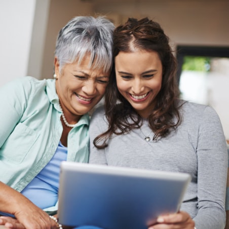 A parent and their adult child laugh while watching a tablet together.
